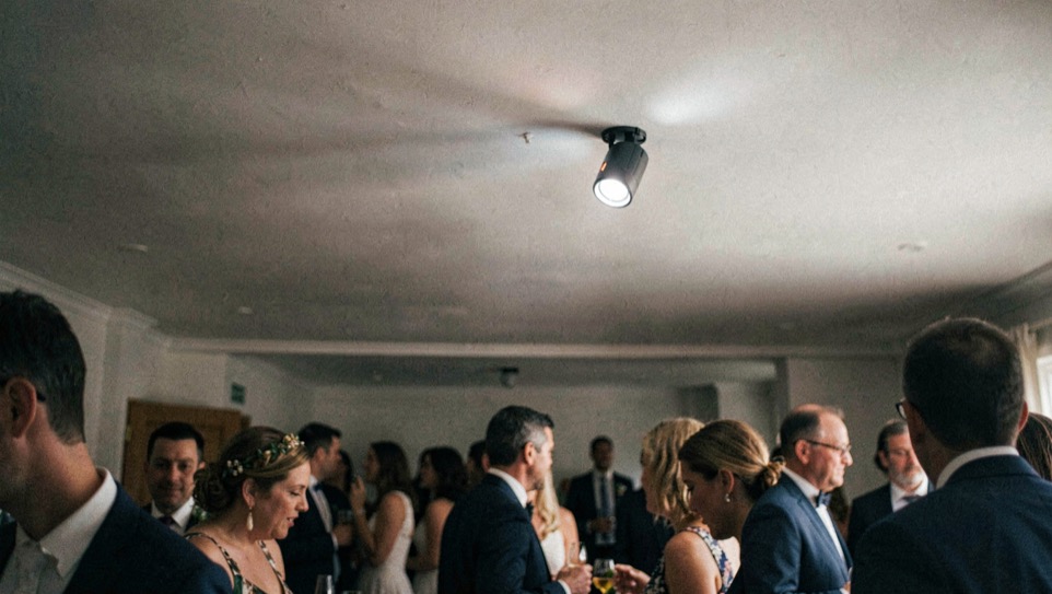 A small speedlight angled toward a pale ceiling while guests move through a dim reception room