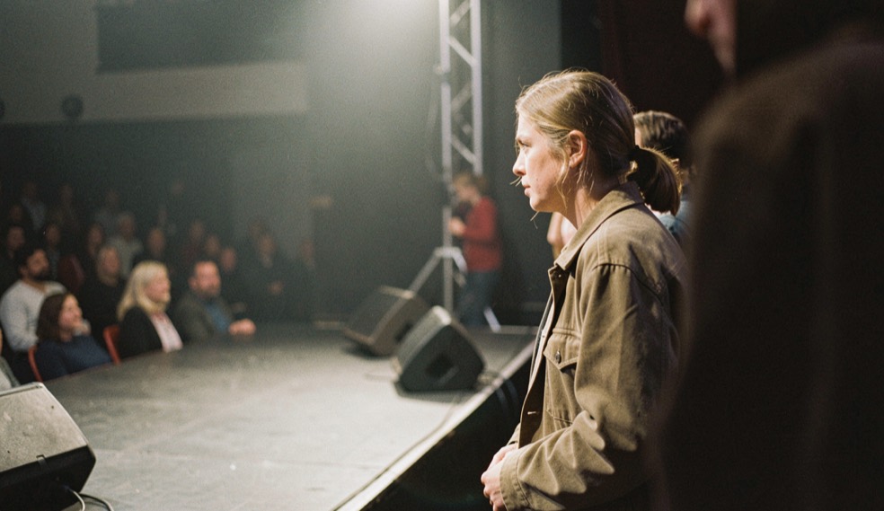 A performer waiting at the edge of a small stage with a soft fill light just out of frame